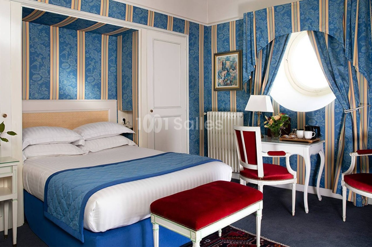 Chambre lumineuse avec lit double, murs à rayures bleues, mobilier classique blanc et rouge, et fenêtre ronde.