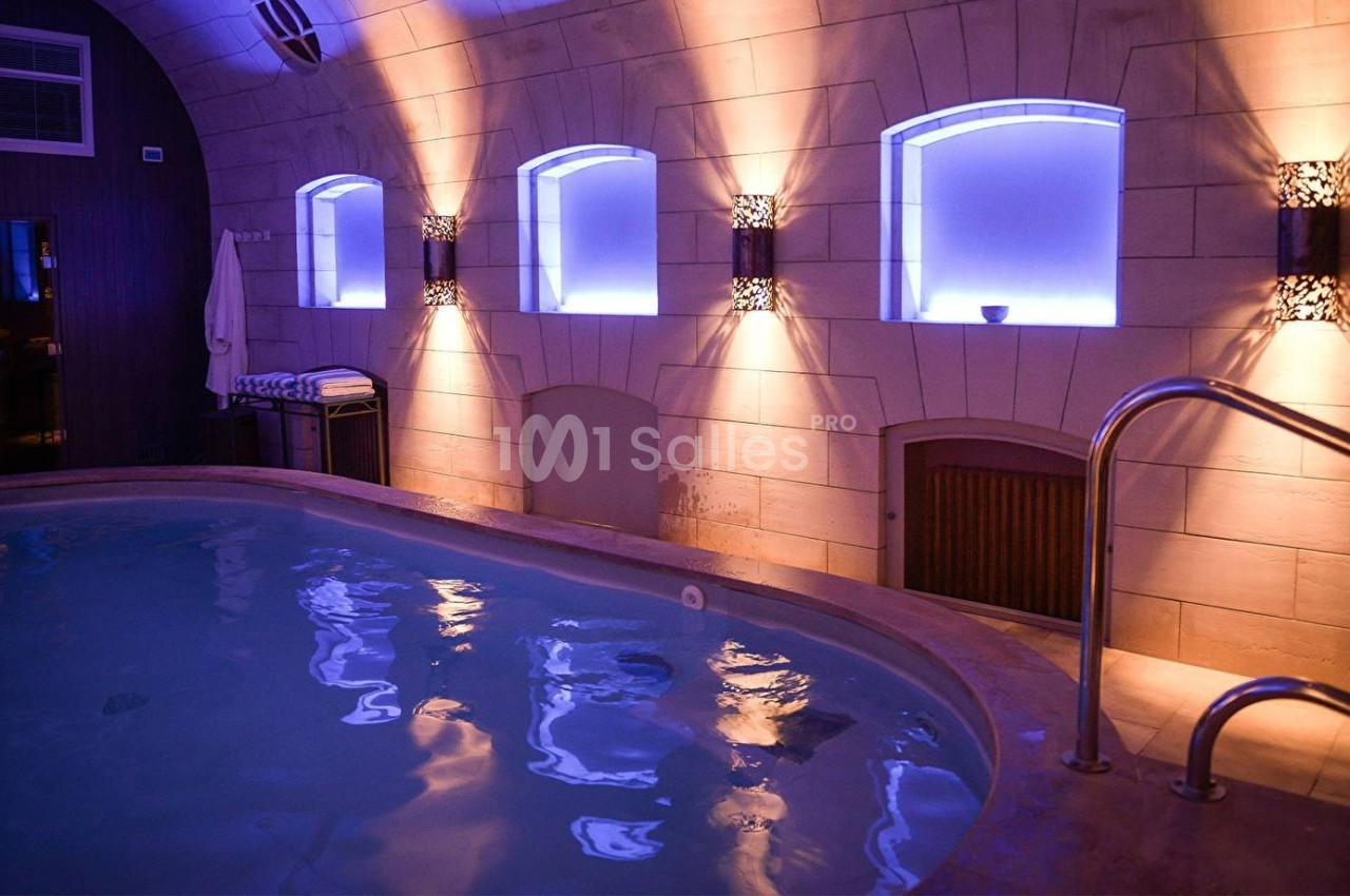 Piscine intérieure éclairée par des lumières tamisées, entourée de murs en pierre avec des fenêtres illuminées.