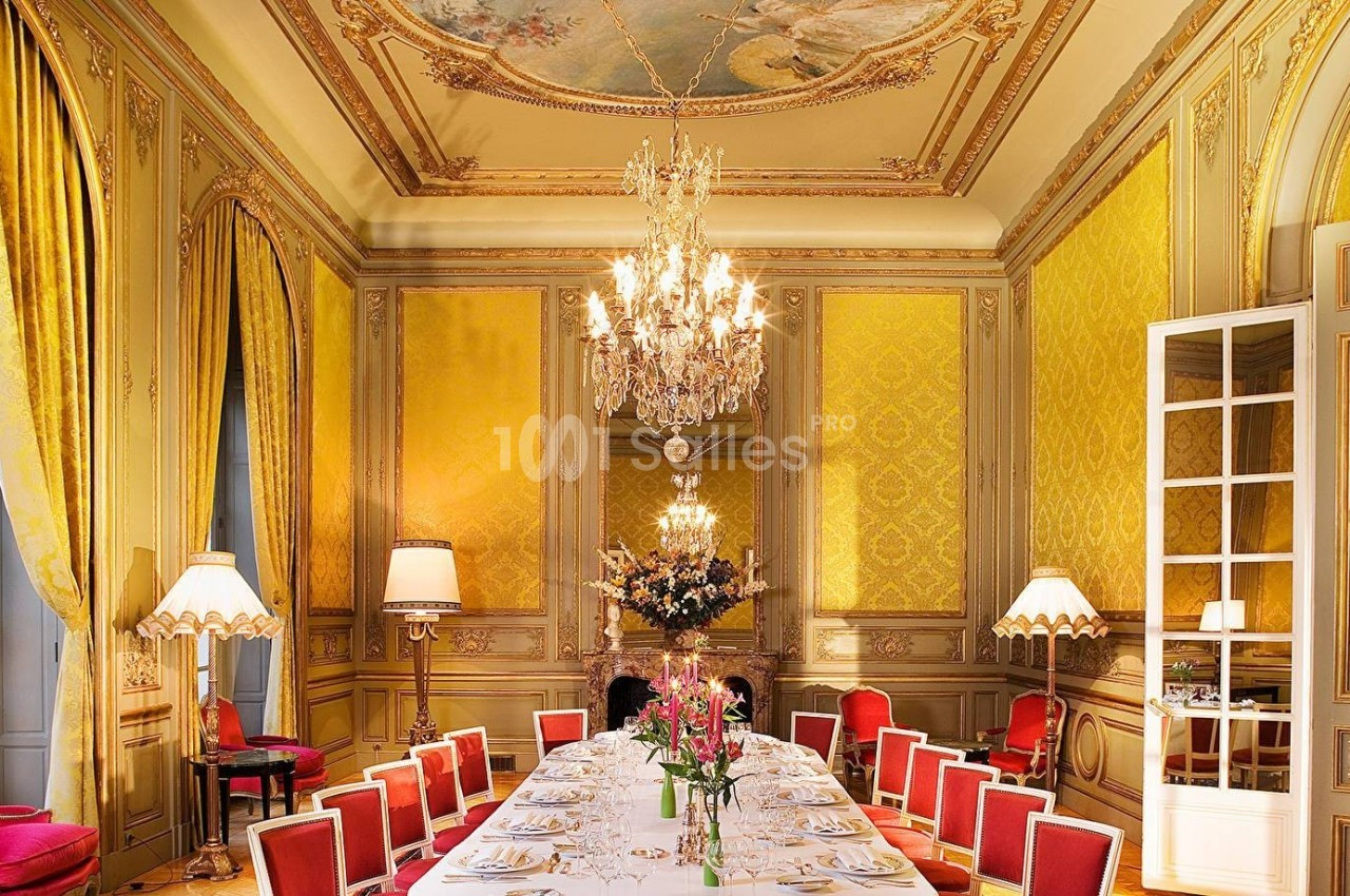 Salle à manger élégante avec un long table dressée, lustres en cristal, murs dorés et plafonds ornés de fresques.