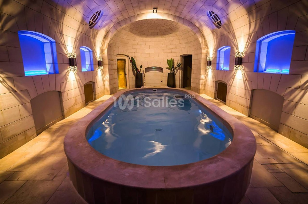 Piscine intérieure ovale dans une pièce voûtée en pierre, éclairée par des lumières bleues et des appliques murales.