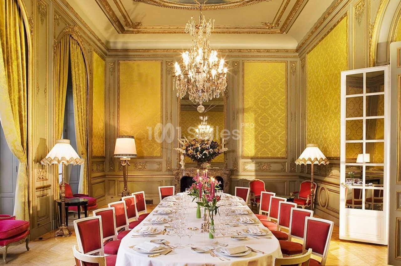 Salle à manger élégante avec table dressée, chaises rouges, murs dorés, lustres et grandes fenêtres avec rideaux jaunes.