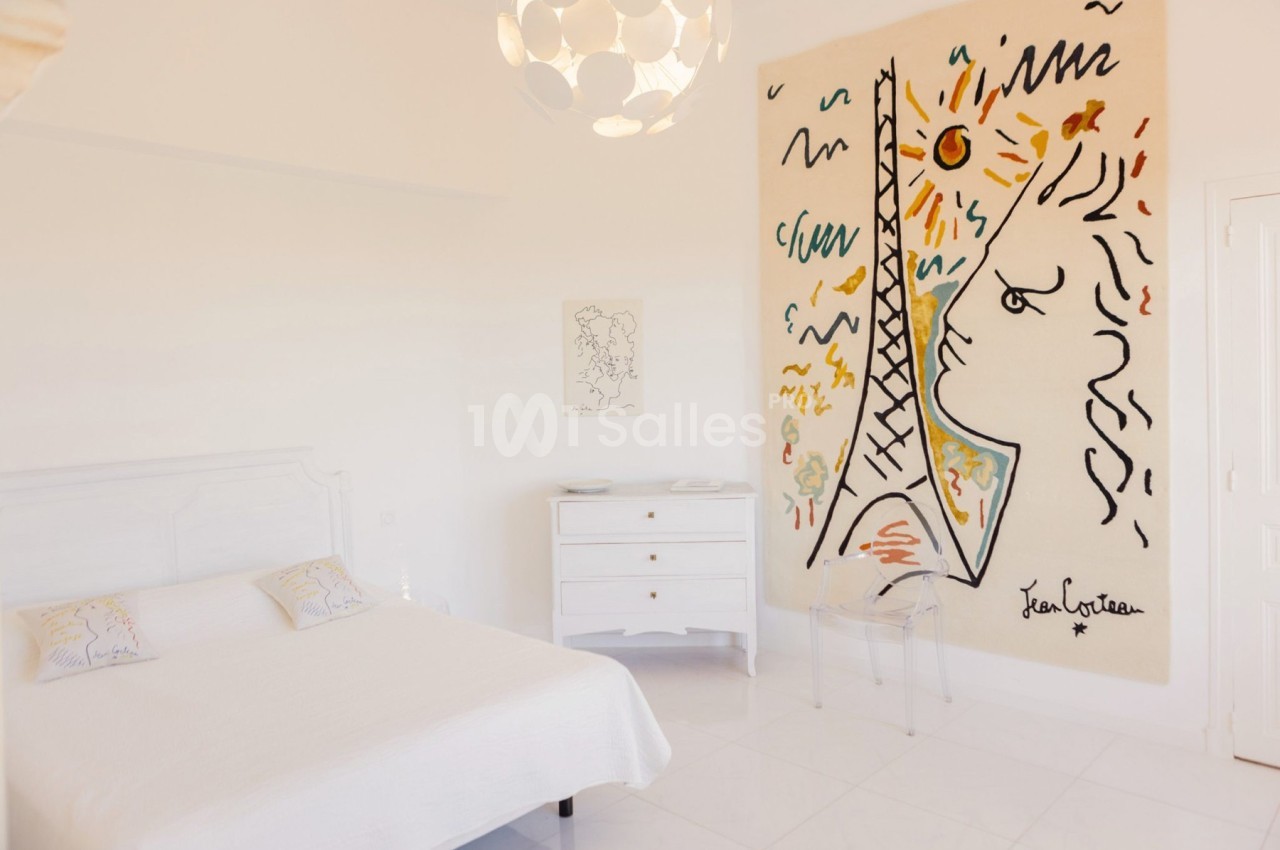 Chambre lumineuse avec un lit double, une commode blanche et une grande œuvre murale colorée représentant une tour et un…