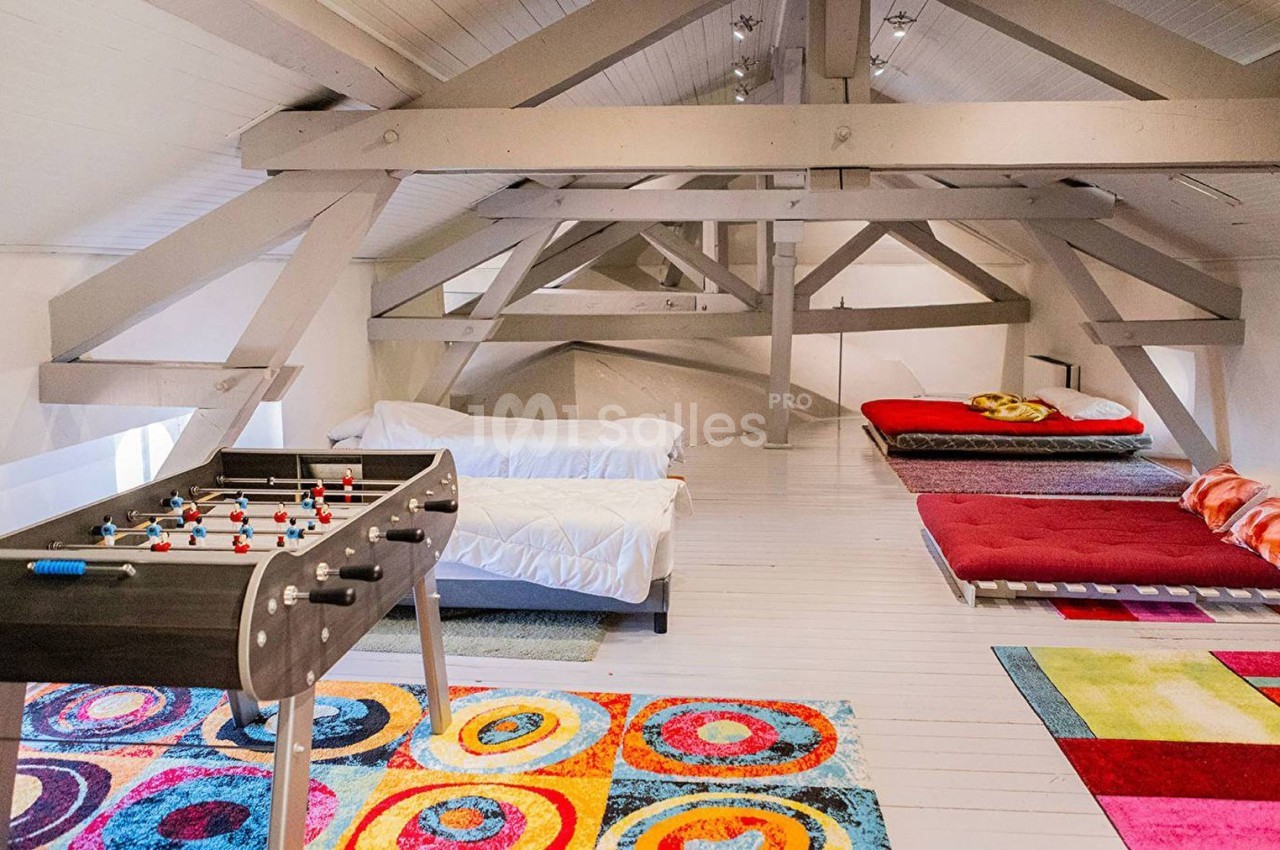 Grenier aménagé avec lits, baby-foot et tapis colorés, sous une charpente apparente en bois peint.