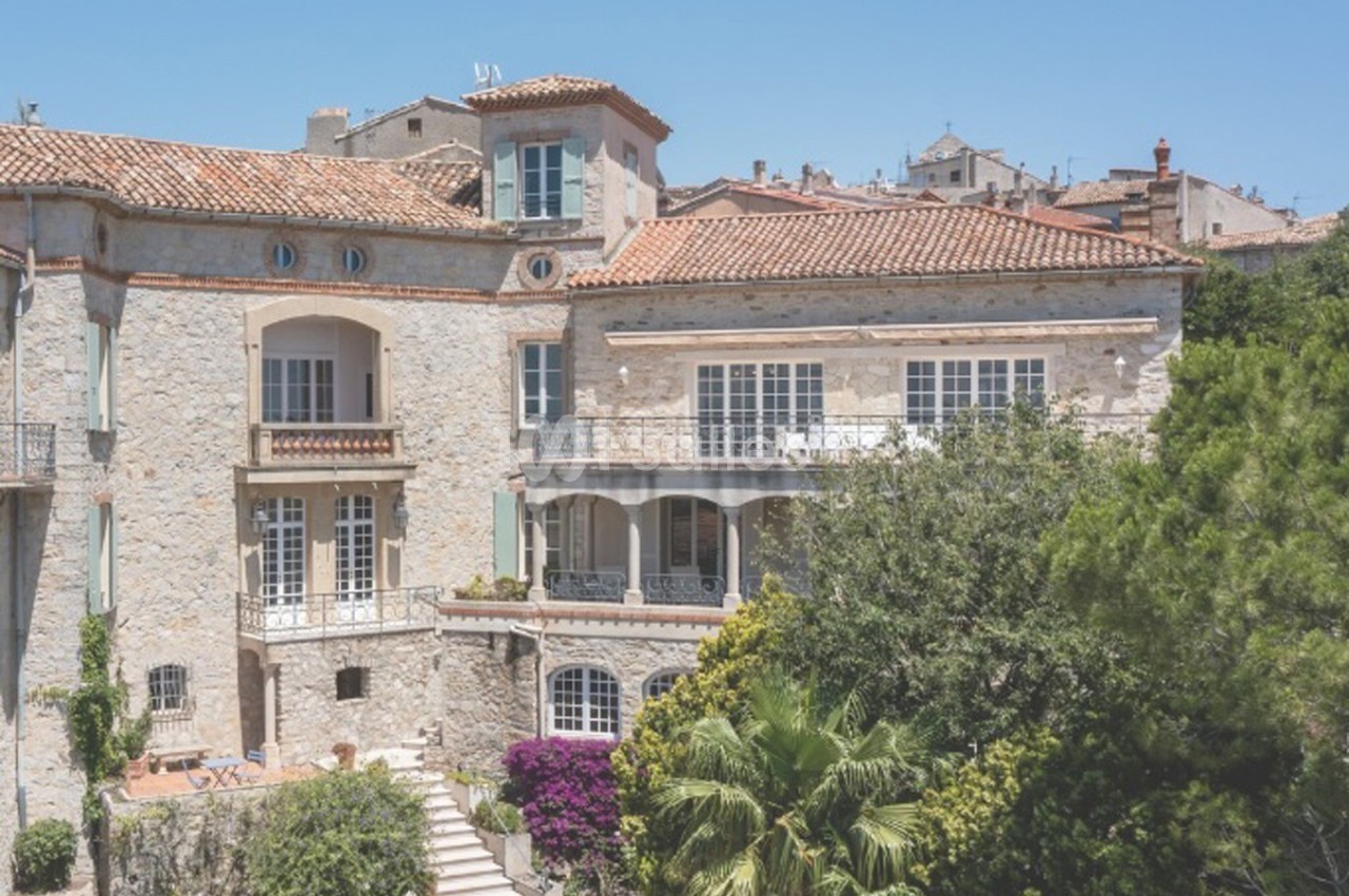 Façade d'une grande maison en pierre avec balcons, entourée de végétation et baignée de lumière sous un ciel bleu.