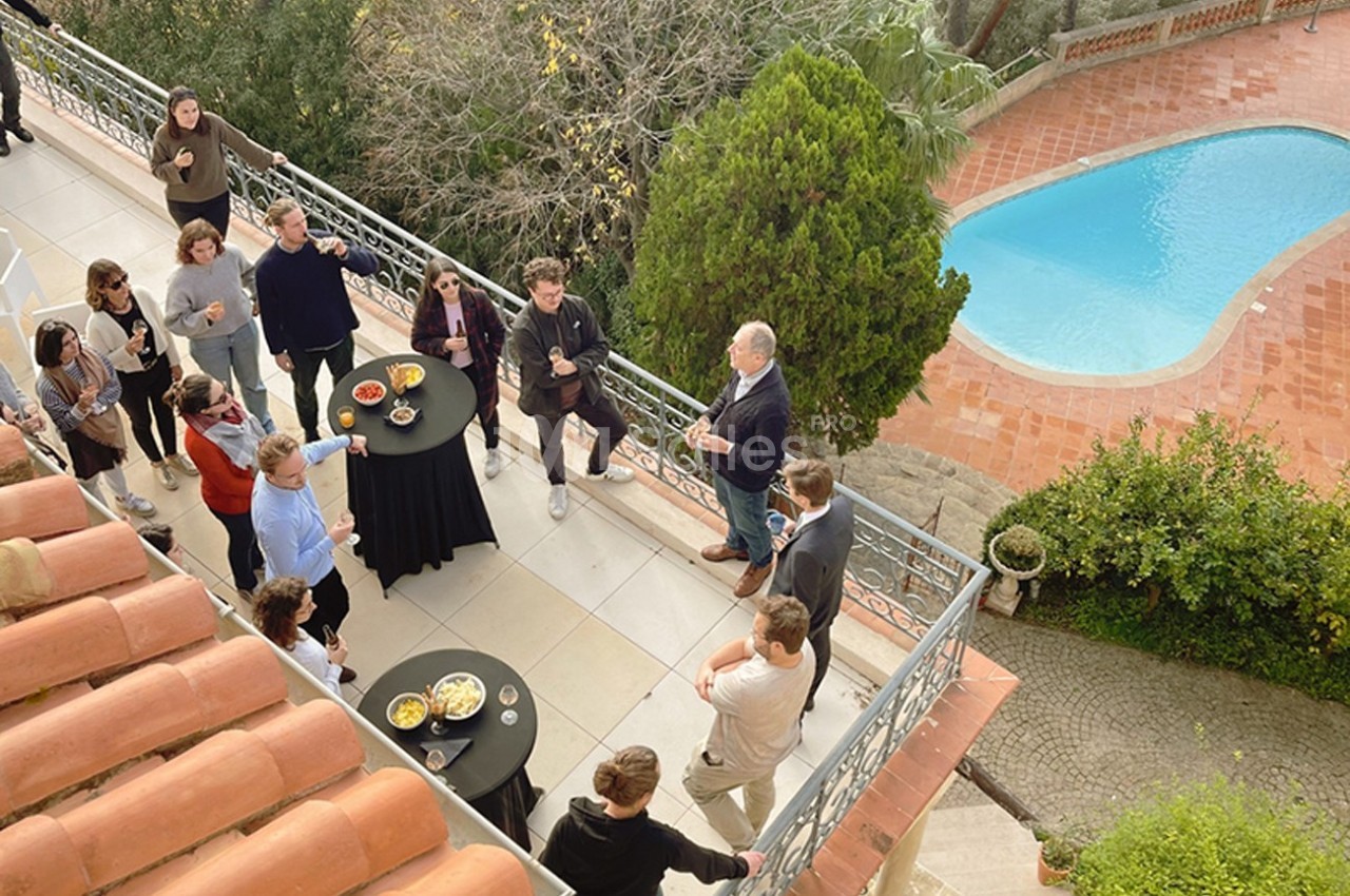 Groupe de personnes discutant autour de tables hautes sur une terrasse surplombant une piscine.