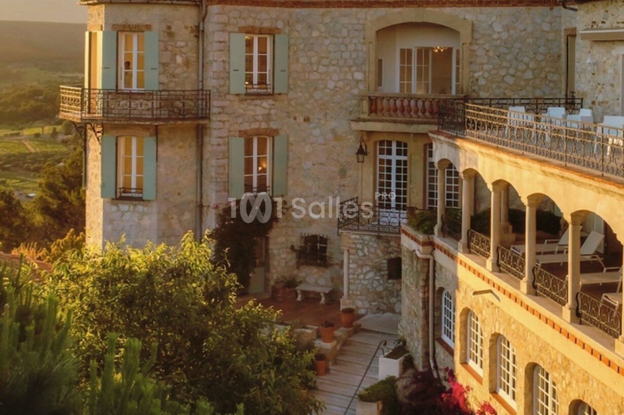 Façade en pierre d'un bâtiment avec balcons et terrasses, entouré de végétation sous une lumière dorée.