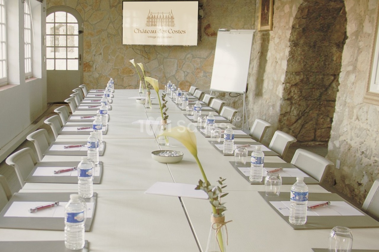 Salle de réunion avec une longue table dressée, équipée de blocs-notes, bouteilles d'eau et fleurs décoratives.