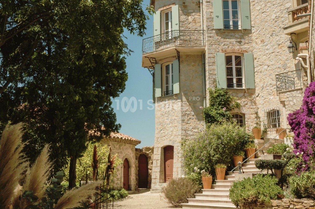 Façade en pierre d'une maison méditerranéenne avec volets verts, escaliers fleuris et végétation environnante.