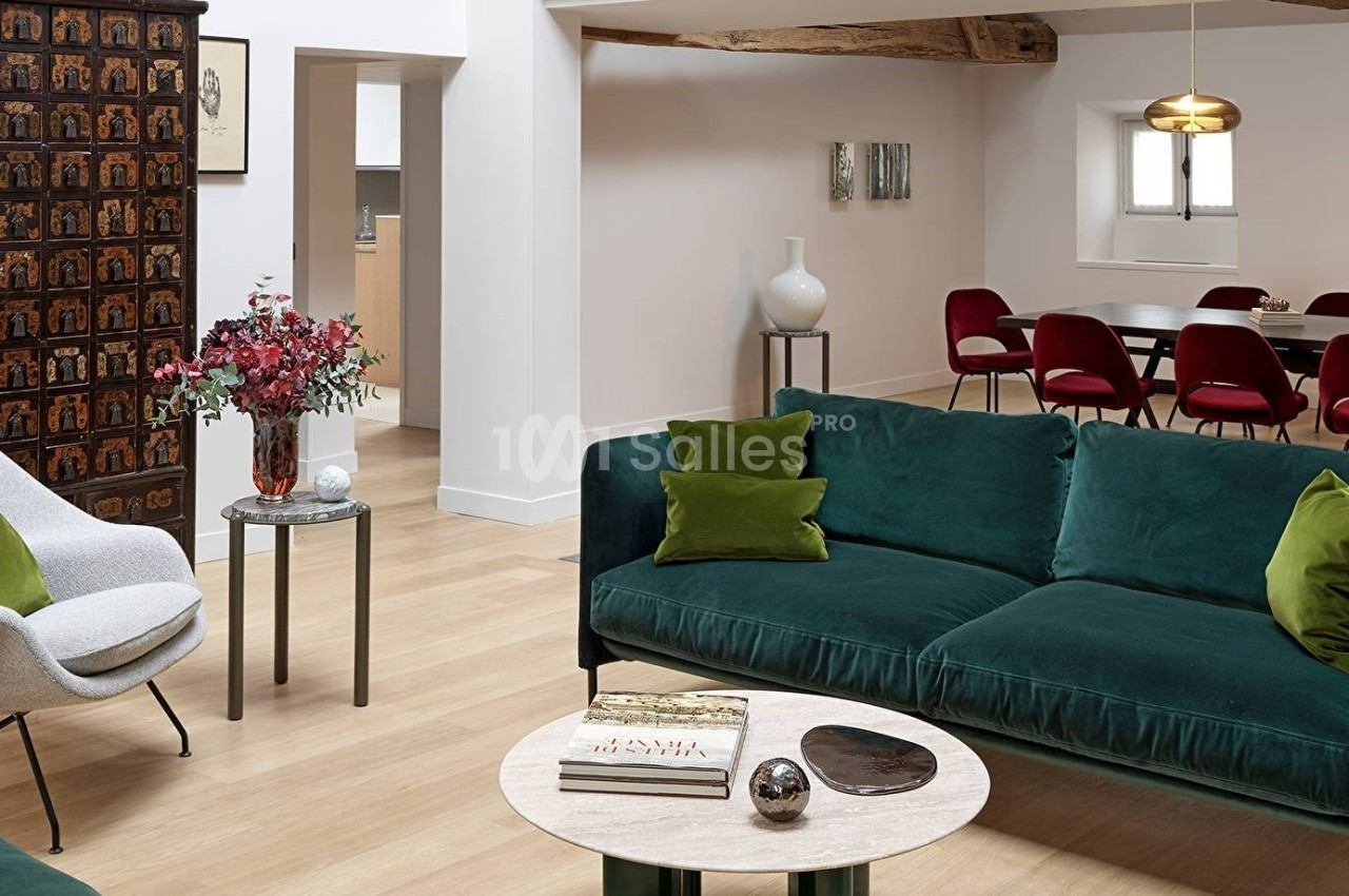 Salon lumineux avec canapé vert, fauteuil blanc, table basse, et salle à manger avec chaises rouges en arrière-plan.