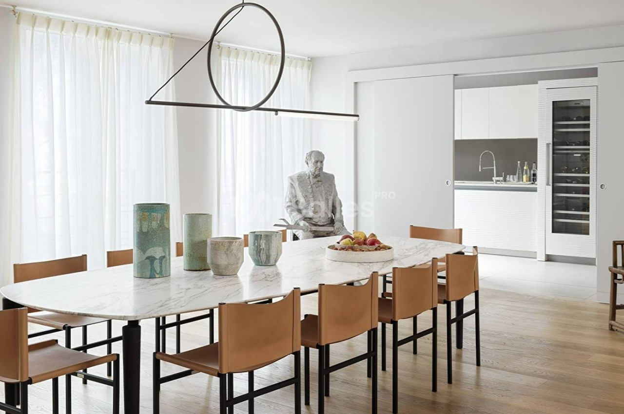 Salle à manger moderne avec table ovale en marbre, chaises en cuir, suspension design et cuisine ouverte en arrière-plan.