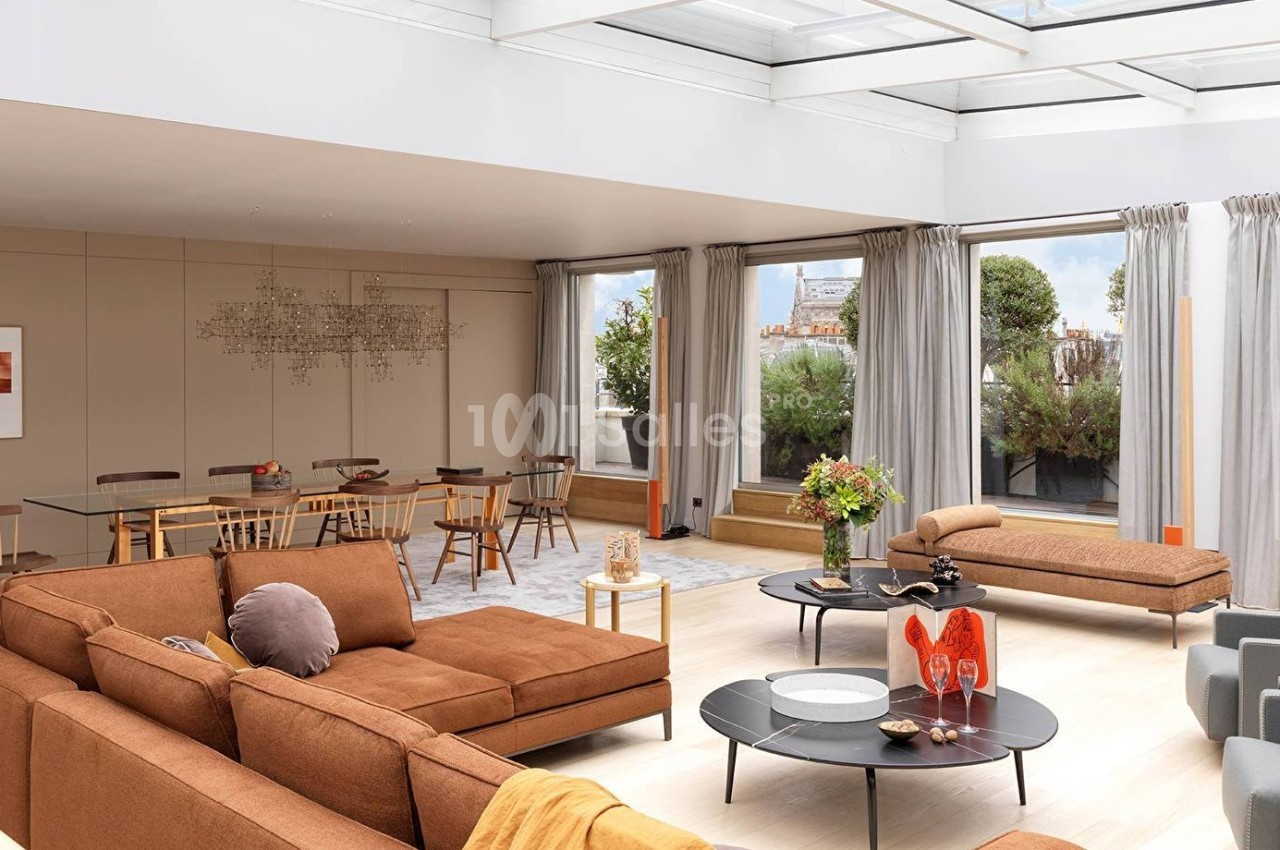 Salon lumineux avec canapé modulable brun, table à manger en verre, grandes baies vitrées et terrasse arborée.