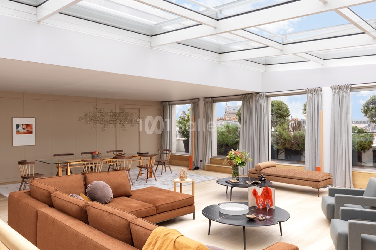 Salon lumineux avec toit vitré, mobilier moderne aux tons chauds, table à manger en arrière-plan et vue sur un jardin.