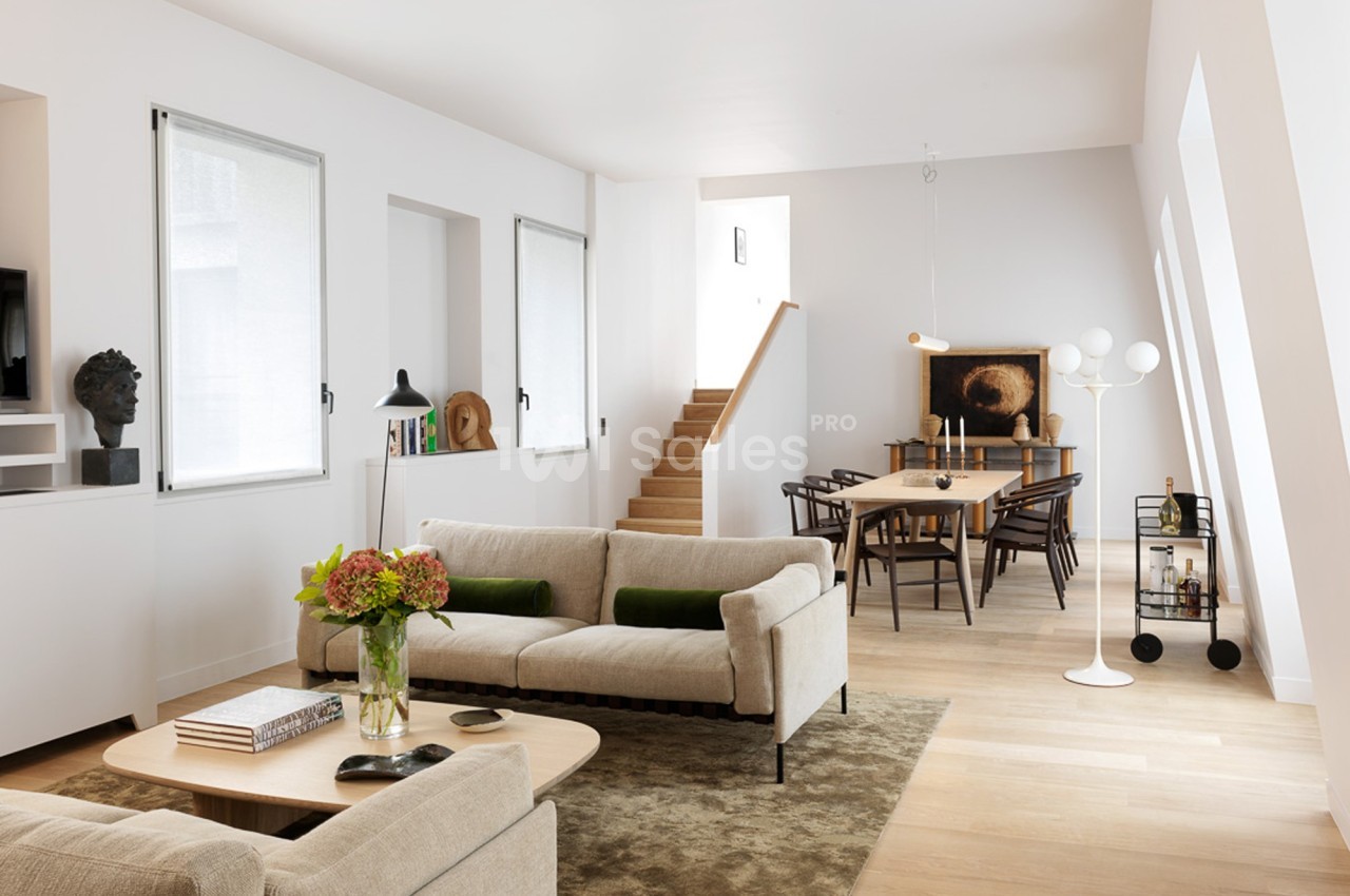 Salon lumineux avec canapé beige, table basse en bois, salle à manger attenante et escalier en arrière-plan.