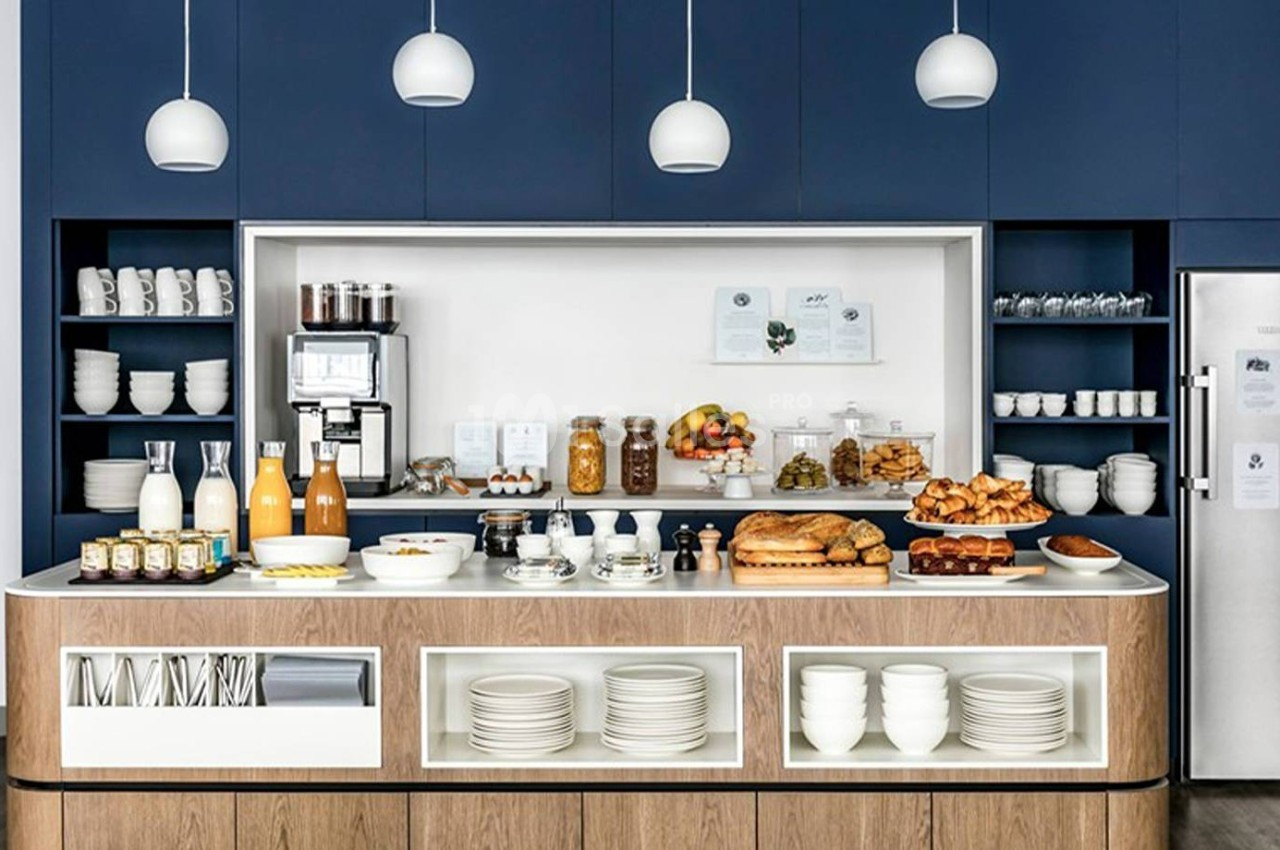 Buffet de petit-déjeuner avec boissons, viennoiseries, fruits et vaisselle, dans une cuisine moderne aux tons bleus et bois.