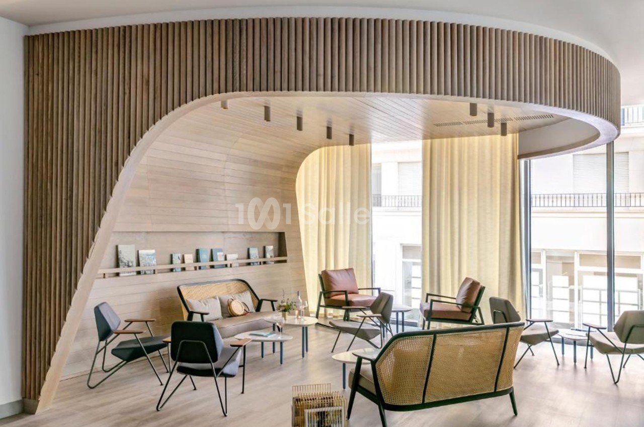 Espace lounge moderne avec mobilier en bois et rotin, éclairé par de grandes baies vitrées et des rideaux légers.