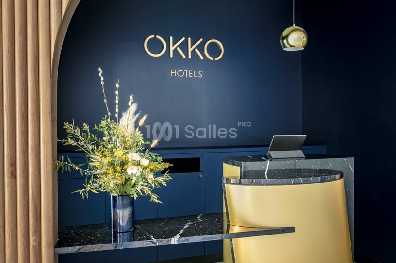 Accueil d’un hôtel avec un comptoir moderne en marbre, une décoration florale et un mur bleu portant le logo ’OKKO HOTELS’.