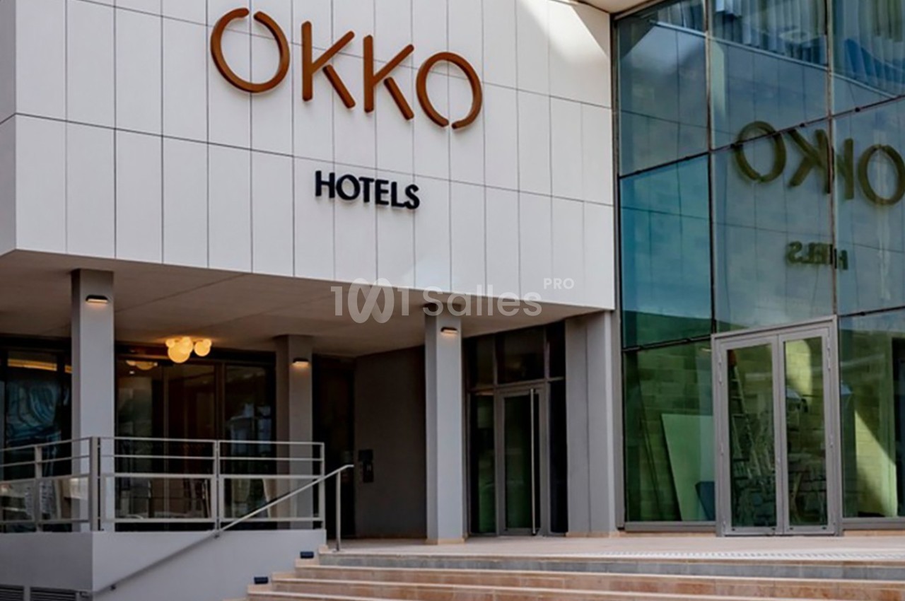 Façade moderne d'un hôtel avec le logo ’OKKO Hotels’, entrée vitrée et escalier menant à la porte principale.