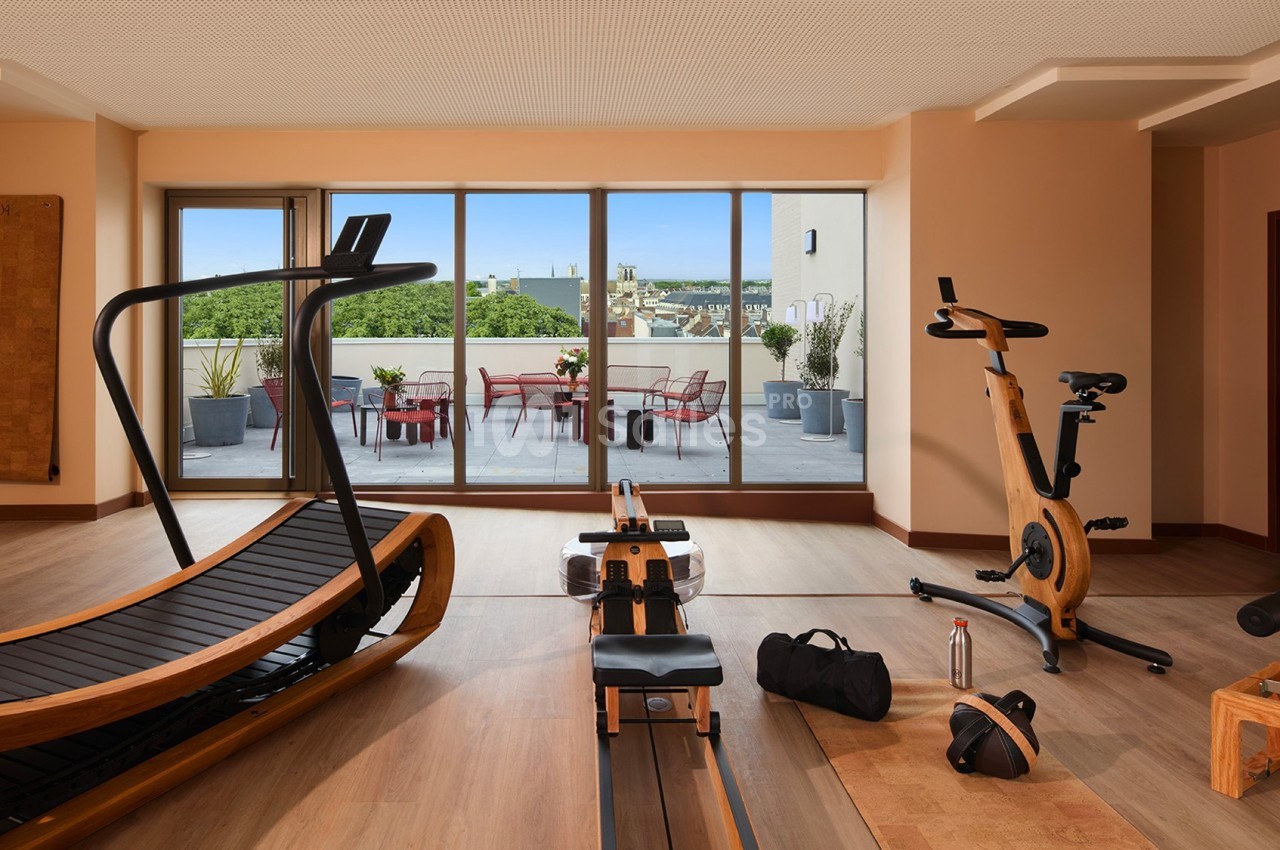 Salle de sport lumineuse avec tapis de course, rameur et vélo d'exercice, donnant sur une terrasse aménagée.