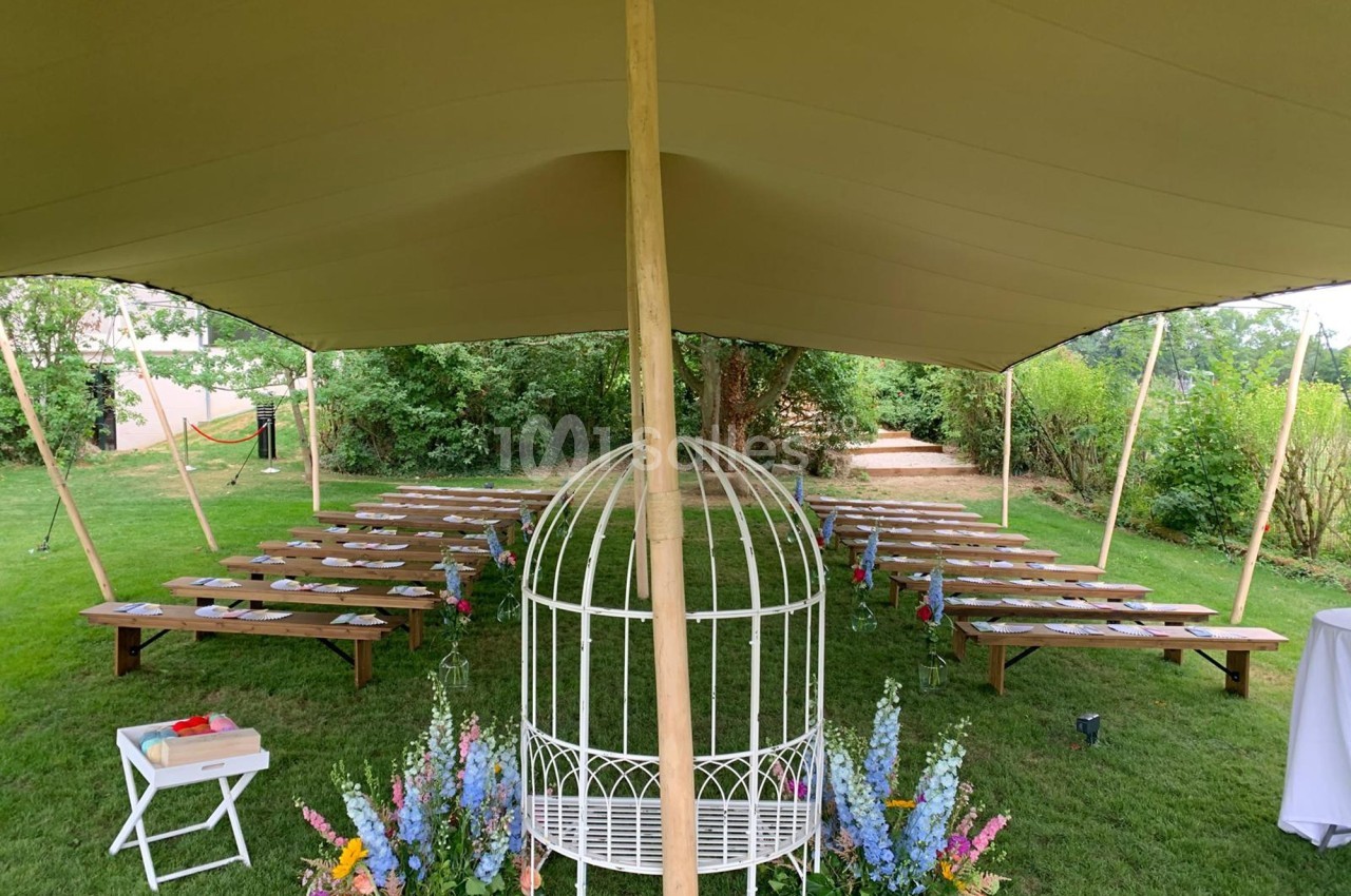 Chaises et bancs disposés sous une tente dans un jardin, avec une arche décorée de fleurs au premier plan.