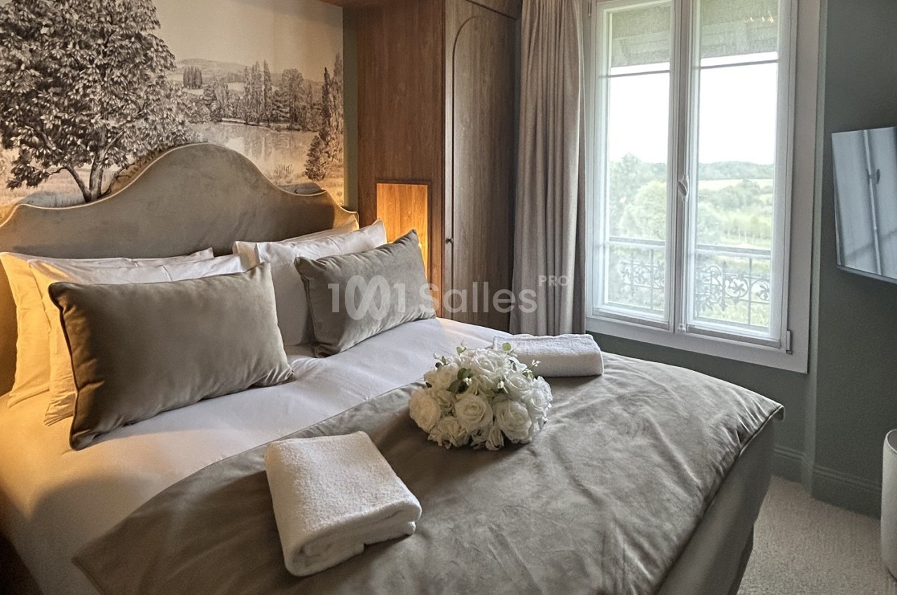 Chambre lumineuse avec lit double, coussins beiges, bouquet de fleurs blanches et vue sur un paysage verdoyant par la…