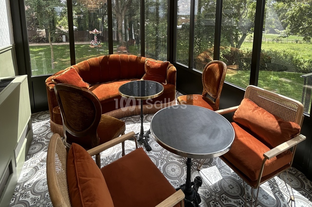 Salon lumineux avec fauteuils et canapé en velours orange, tables rondes et vue sur un jardin verdoyant.