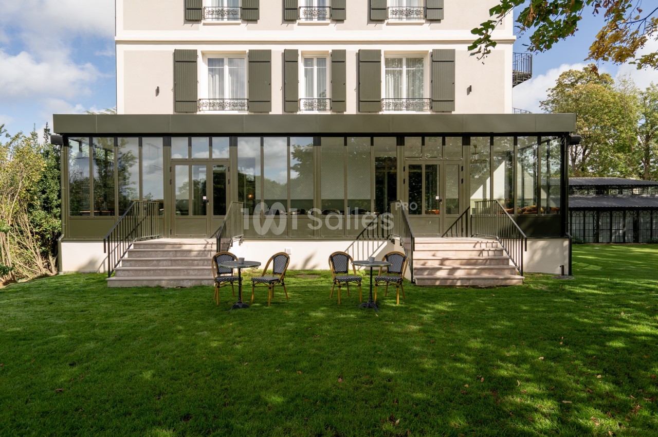 Façade d'un bâtiment avec une véranda vitrée, escaliers et chaises disposées sur une pelouse verte.