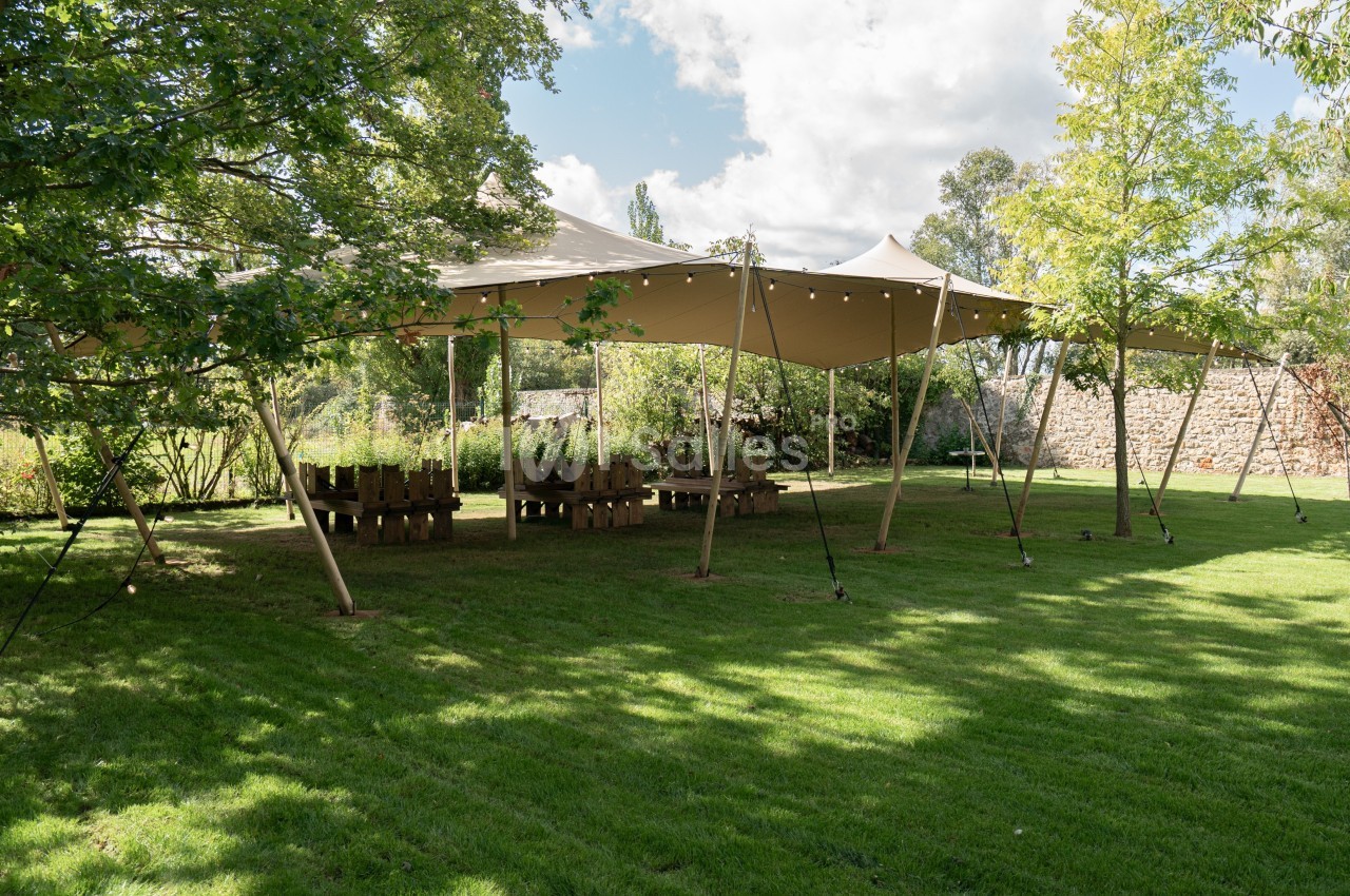 Tente beige montée dans un jardin verdoyant, entourée d'arbres, avec des tables en bois disposées dessous.