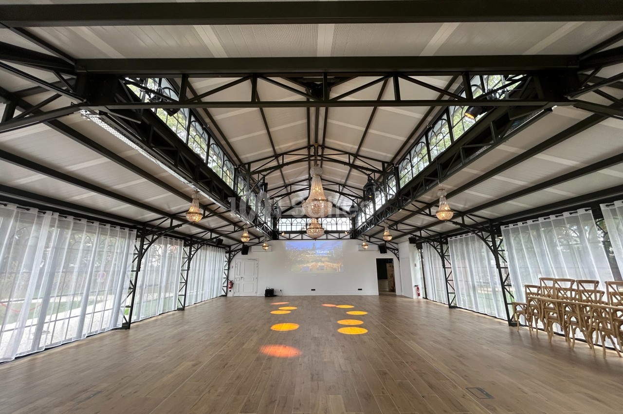 Salle lumineuse avec structure métallique, parquet en bois, lustres suspendus et chaises alignées sur le côté.
