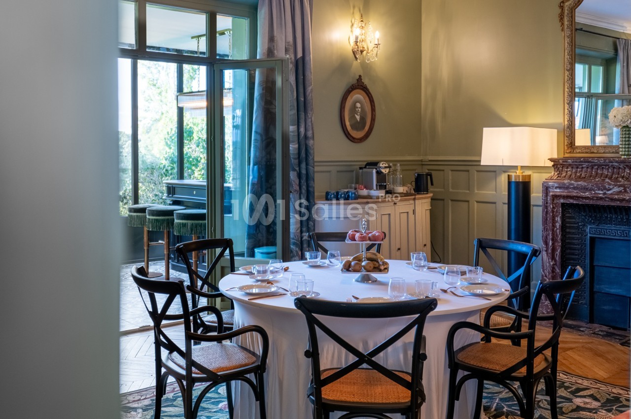 Salle à manger élégante avec table ronde dressée, chaises en bois, buffet et lumière naturelle entrant par une baie vitrée.