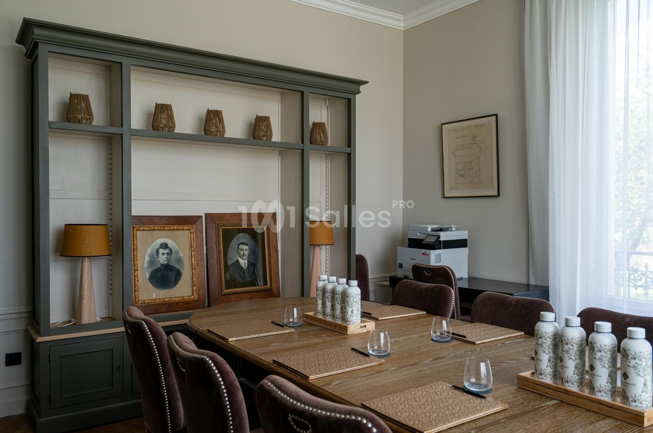 Salle de réunion avec table en bois, chaises, étagère décorative, portraits anciens et une imprimante en arrière-plan.