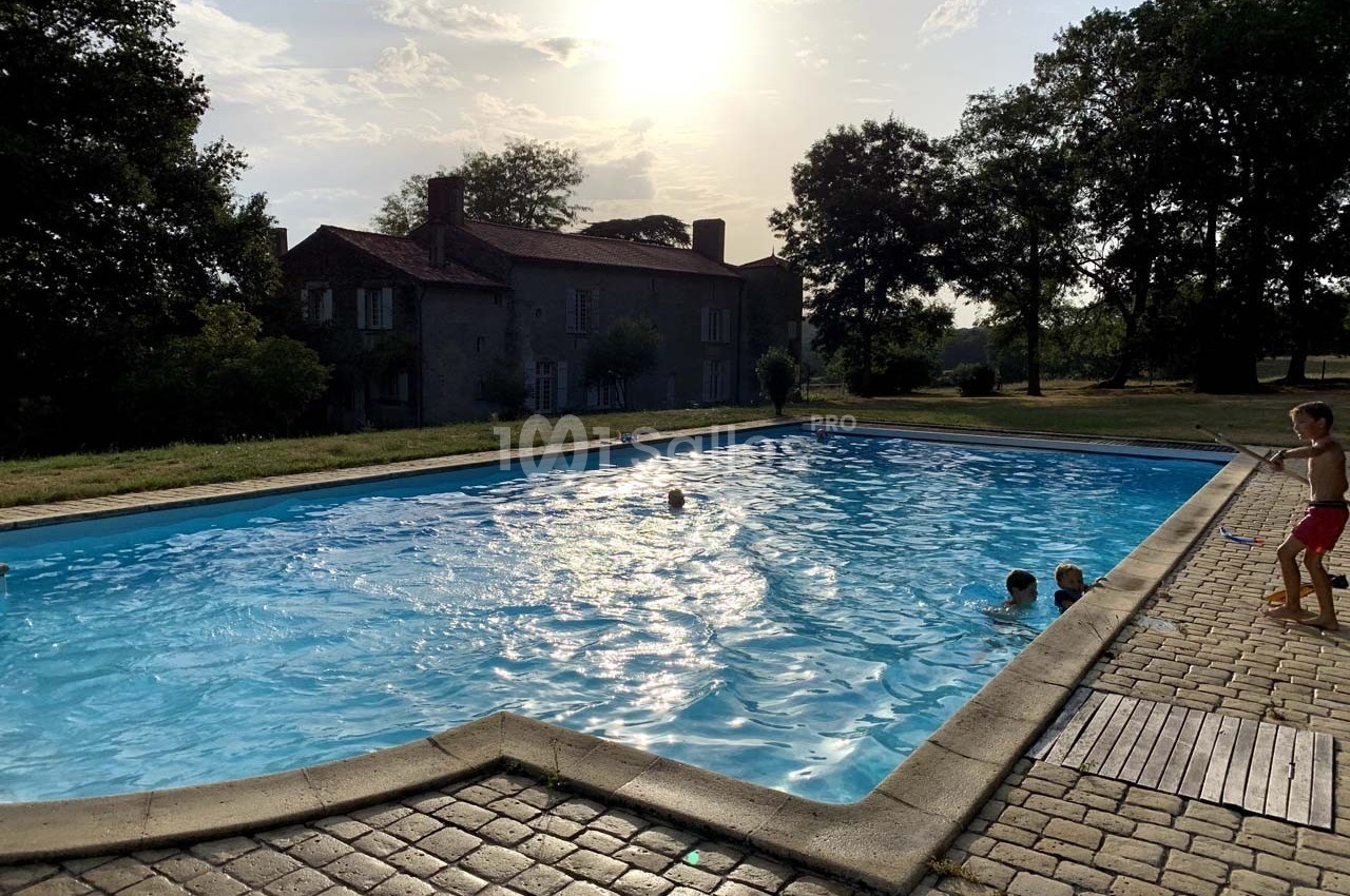 Piscine extérieure entourée de pavés, avec des enfants jouant dans l'eau près d'une maison en pierre au coucher du soleil.