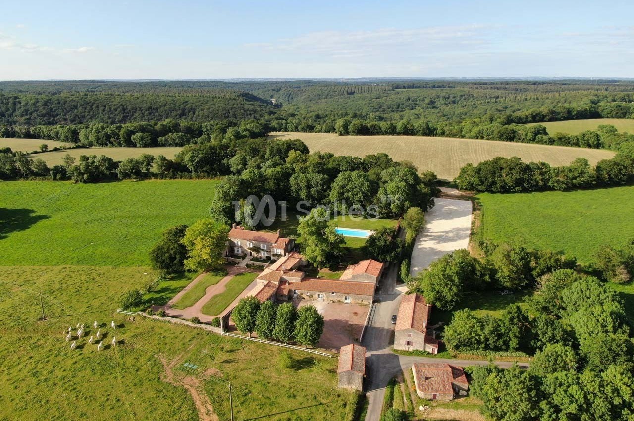 Vue aérienne d'une ferme avec bâtiments en pierre, piscine, champs verdoyants et forêt en arrière-plan.