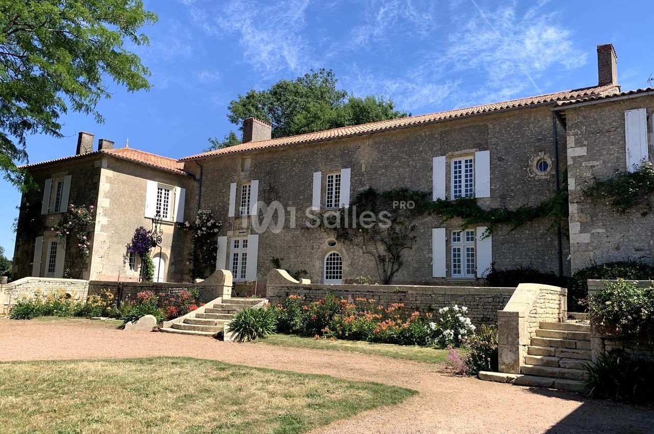 Façade d'une grande maison en pierre avec volets blancs, entourée de jardins fleuris et d'escaliers en pierre.