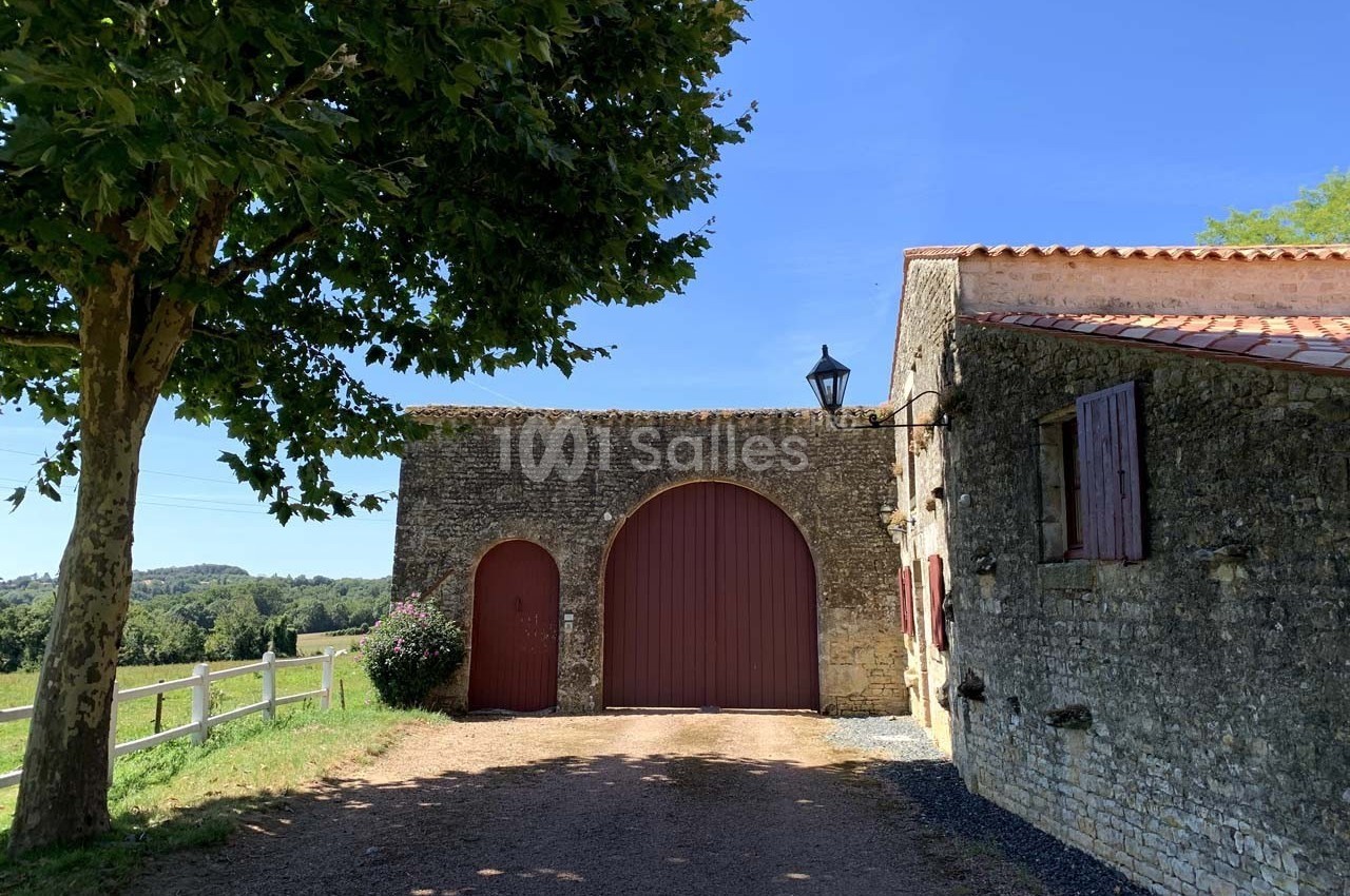 Portail en bois rouge encadré par des murs en pierre, avec un arbre et une vue sur la campagne en arrière-plan.