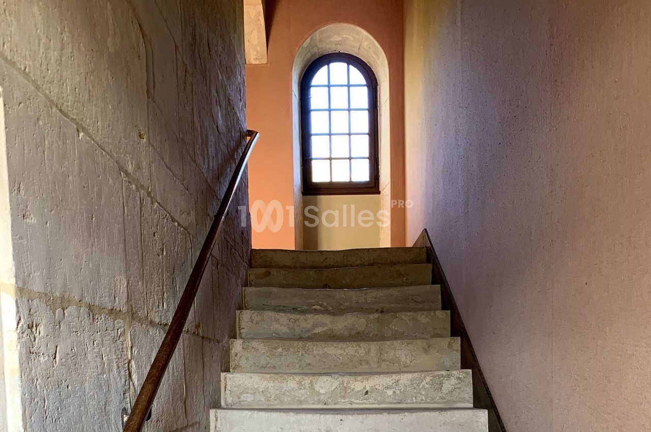 Escalier en pierre avec rampe en métal menant à une fenêtre cintrée laissant passer la lumière naturelle.