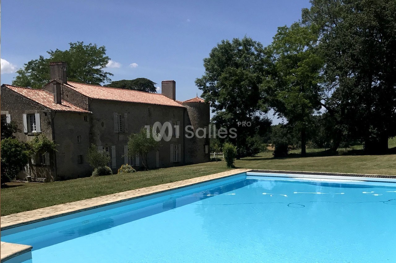 Grande piscine extérieure devant une maison en pierre avec toit en tuiles, entourée d'arbres et de pelouse.
