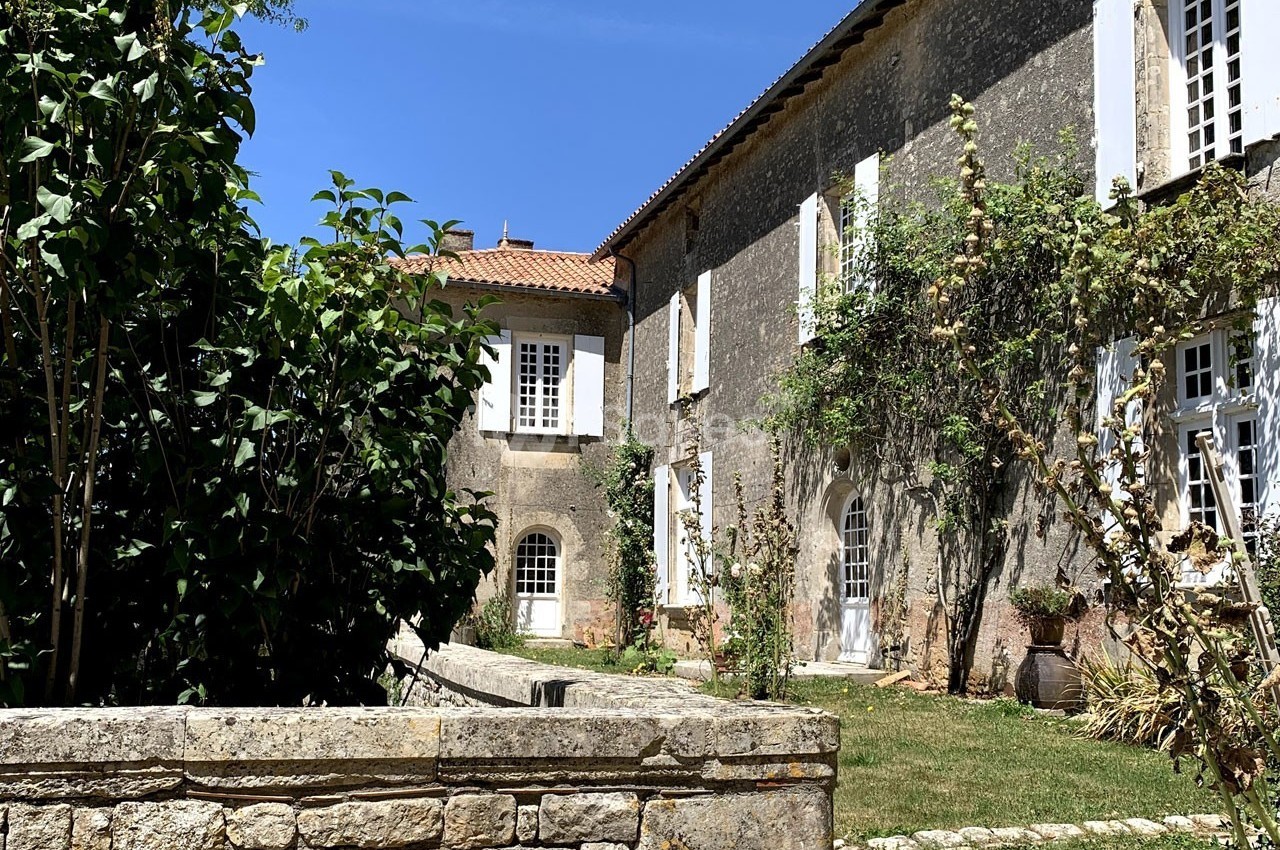 Façade d'une maison en pierre avec volets blancs, entourée de végétation sous un ciel bleu.