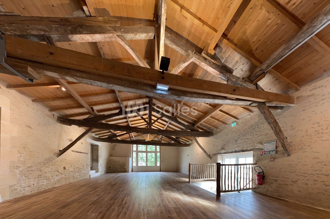 Salle spacieuse avec charpente en bois apparente, murs en pierre et parquet, éclairée par des fenêtres et des lumières…