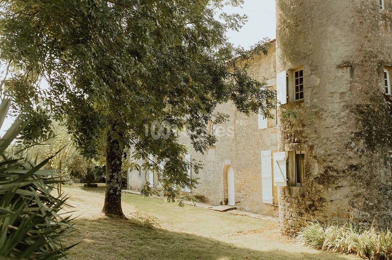 Façade en pierre d'une maison ancienne avec volets blancs, entourée d'arbres et d'un jardin verdoyant.