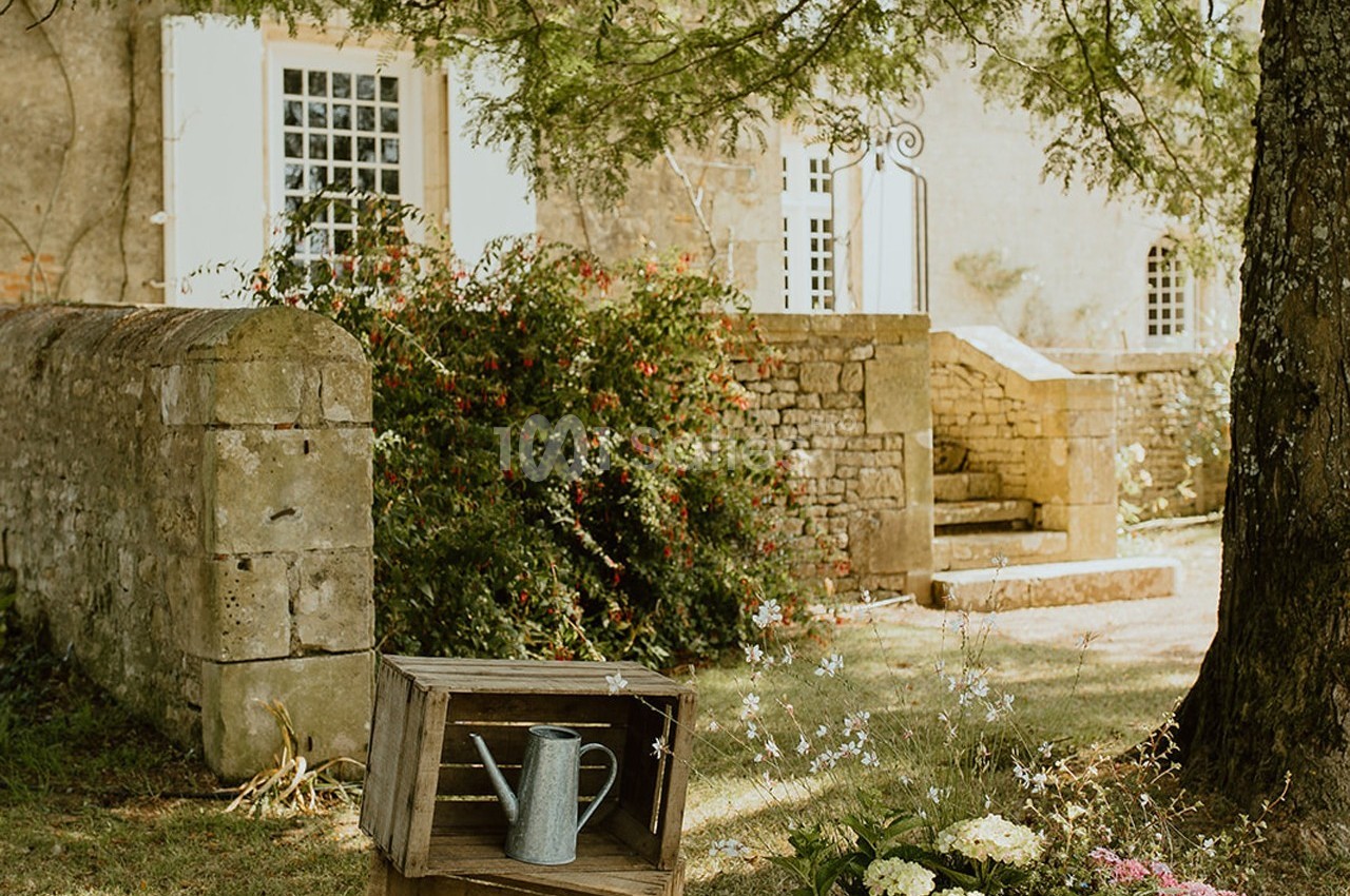 Arrosoir en métal posé sur une caisse en bois dans un jardin fleuri devant une maison en pierre.
