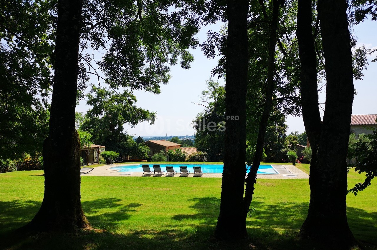 Piscine extérieure entourée de chaises longues, située dans un jardin verdoyant avec des arbres en premier plan.