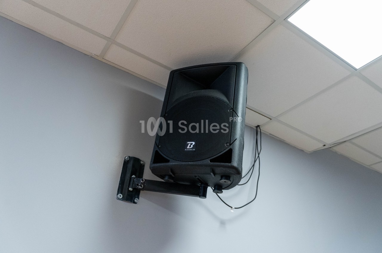 Haut-parleur noir monté sur un mur gris, fixé à un support métallique sous un plafond à dalles blanches.