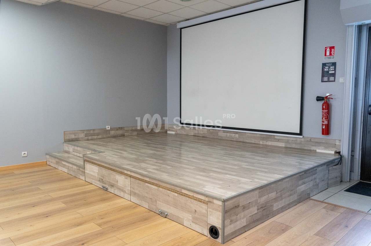 Petite scène en bois clair avec écran de projection blanc en arrière-plan, dans une salle au sol en parquet.