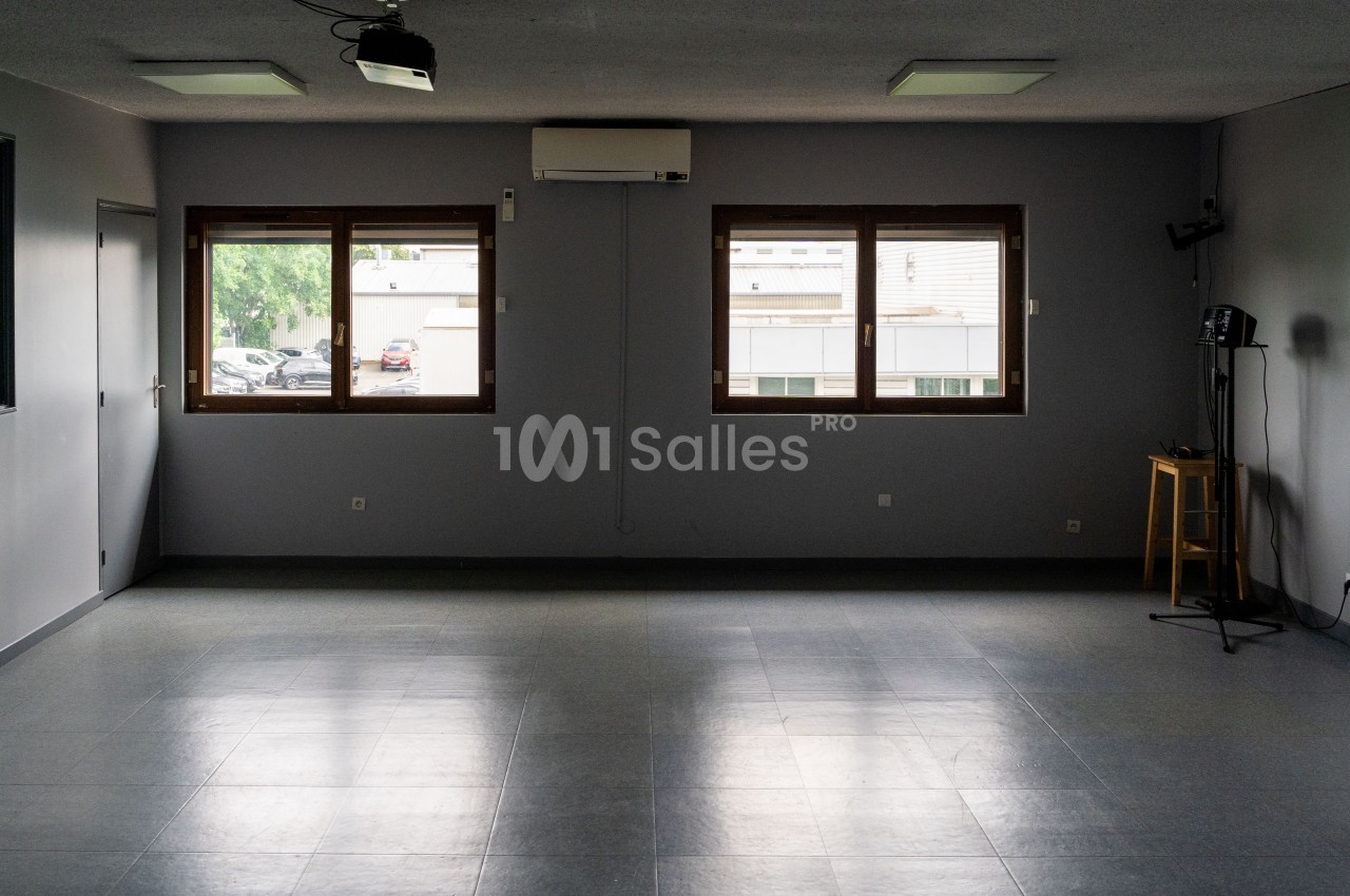 Salle vide avec sol carrelé, murs gris, trois fenêtres laissant entrer la lumière naturelle et quelques équipements.