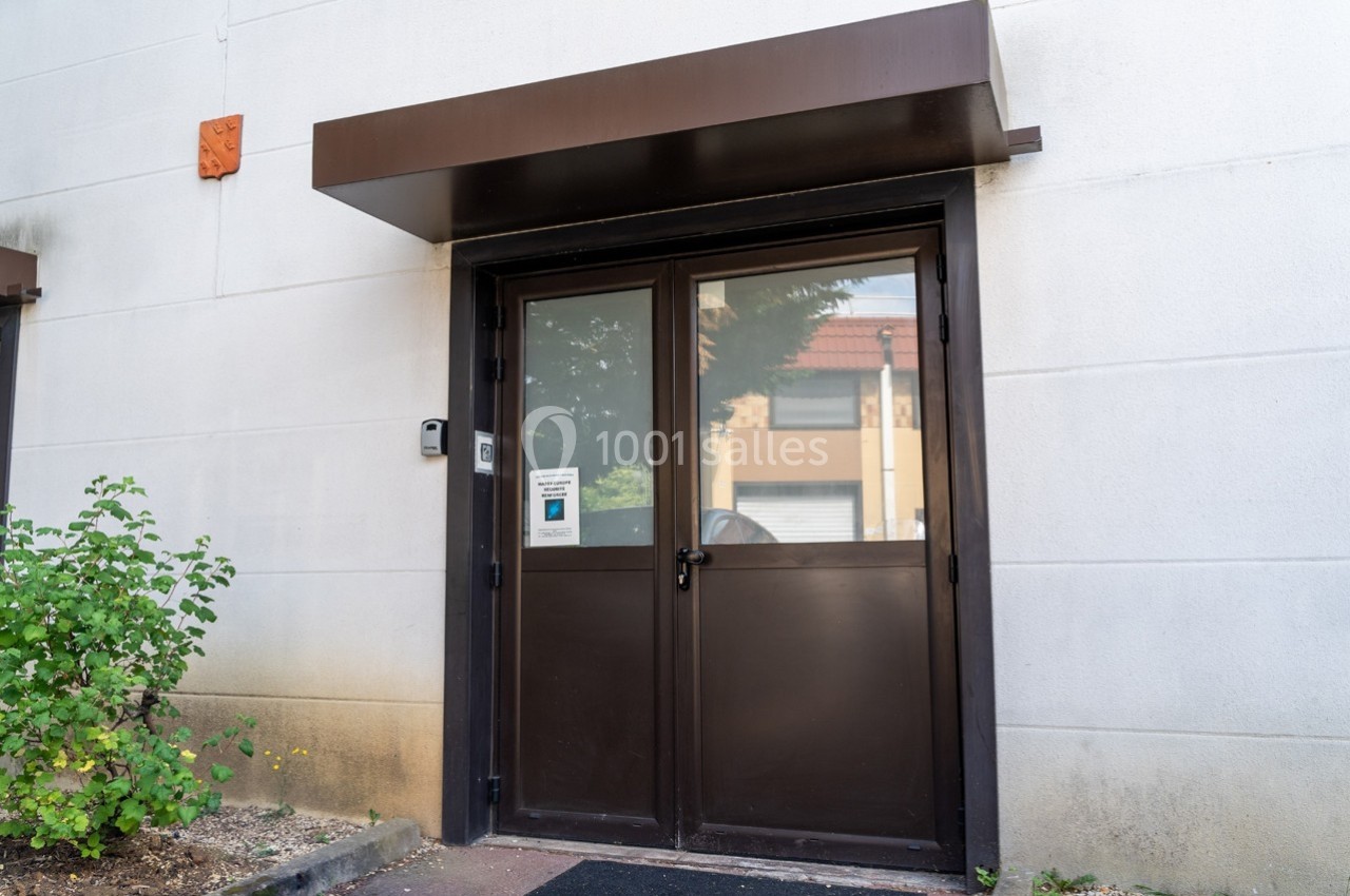 Porte d'entrée vitrée en métal sombre avec auvent, située sur une façade blanche, entourée de végétation.