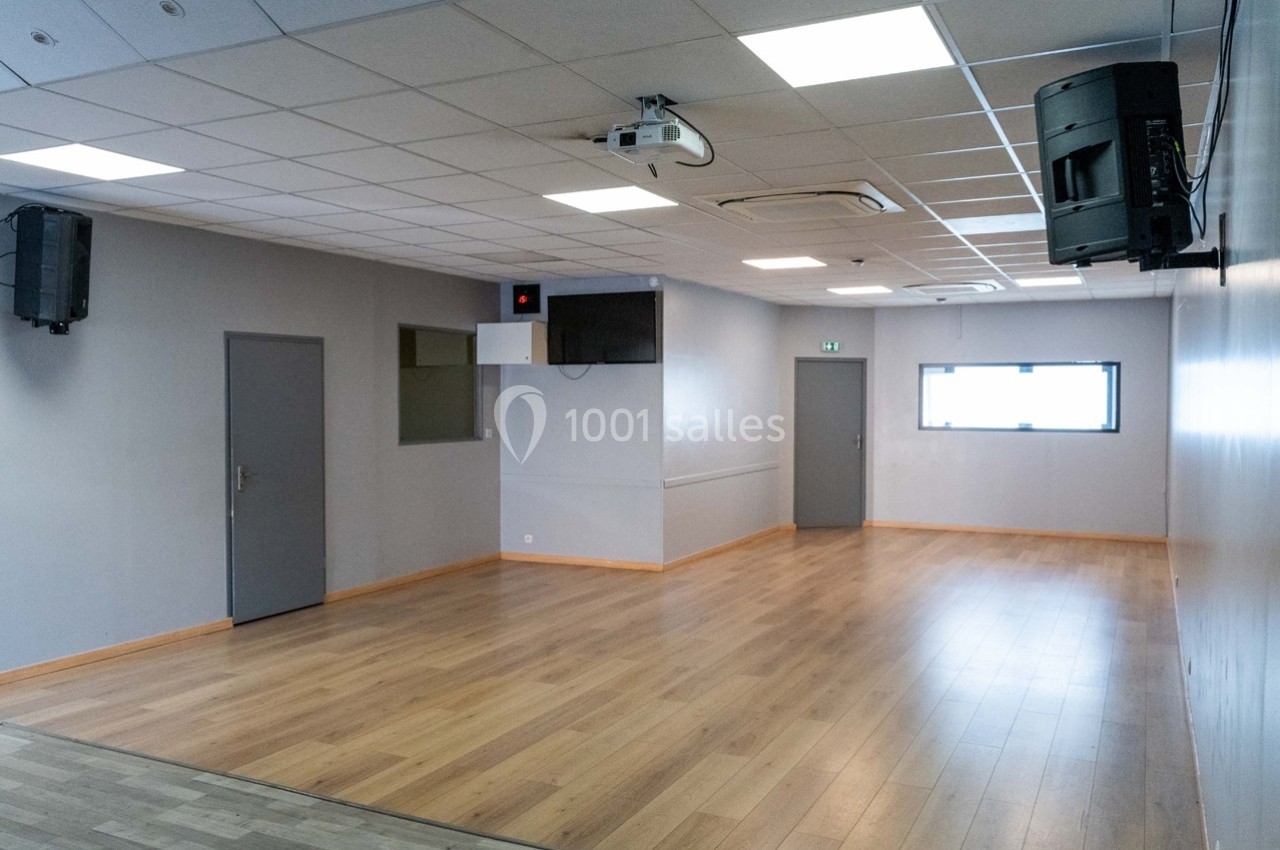Salle vide avec parquet, murs clairs, plafond à dalles, éclairage fluorescent, fenêtres et équipements audiovisuels.