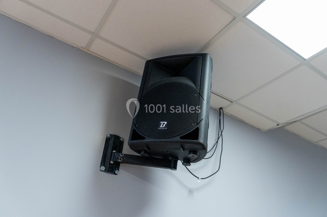 Haut-parleur noir fixé au mur sous un plafond à dalles blanches, avec câbles visibles.
