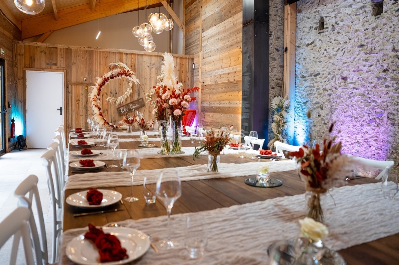 Salle décorée pour un événement, avec longues tables en bois, nappes blanches, fleurs séchées et éclairage chaleureux.