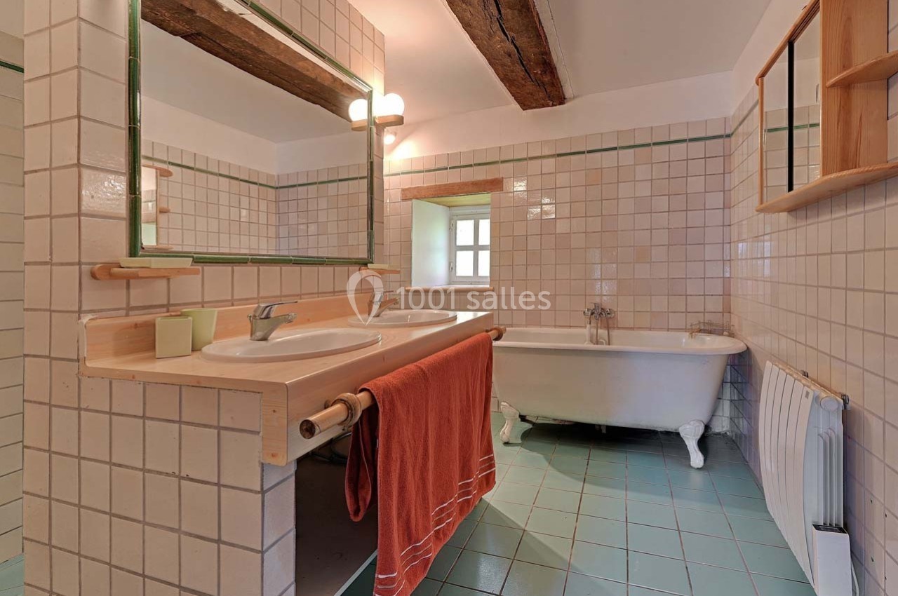 Salle de bain avec carrelage beige et bleu, double vasque, baignoire sur pieds et poutre apparente au plafond.