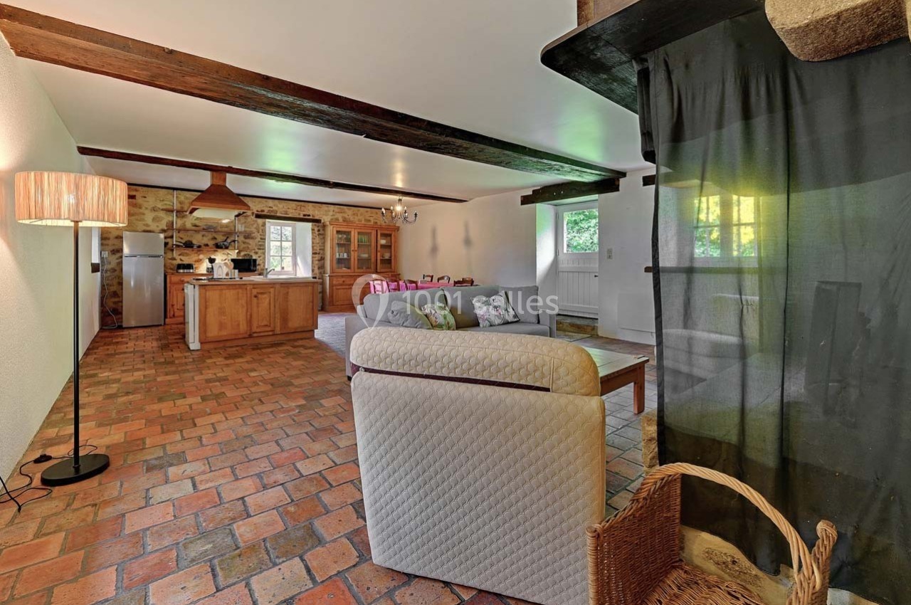 Salon spacieux avec sol en terre cuite, poutres apparentes, canapé, cuisine ouverte et lumière naturelle.